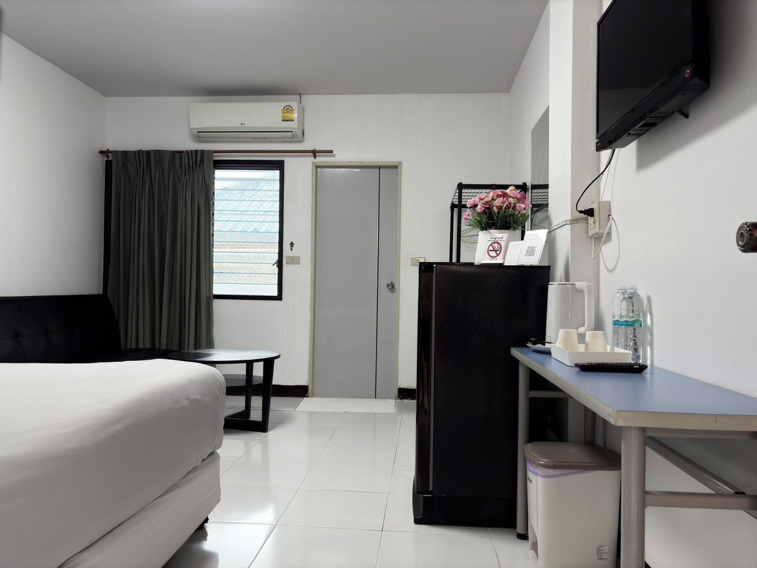 A hotel room with a bed, a desk, a refrigerator, a trash bin, and a television mounted on the wall.