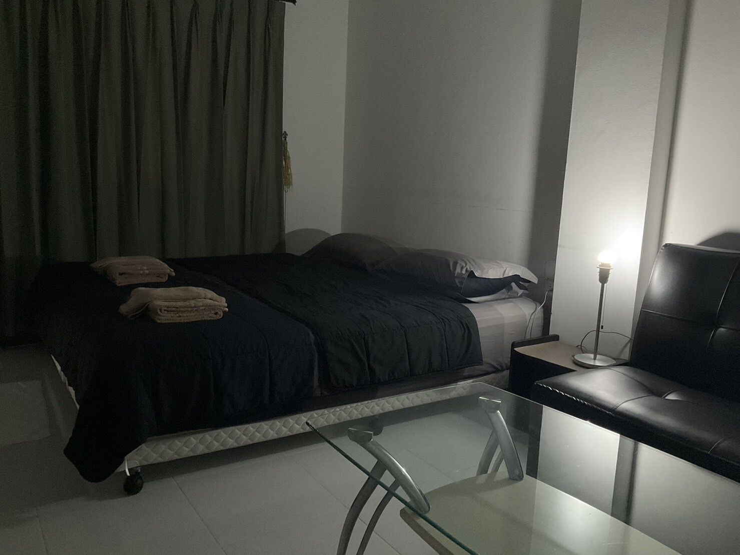 A bedroom with a bed, pillows, a bedside table, a lamp, and a glass table with chairs.