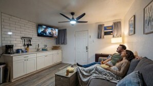 A living room with a sofa, a television, a ceiling fan, and a kitchen area with cabinets and a coffee maker. - Cozy Oceanview Studio on Galveston’s East End (Galveston)