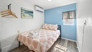 Ein Schlafzimmer mit Bett, Kleiderstange, Klimaanlage, einem Gemälde und einem Fenster mit Vorhängen.