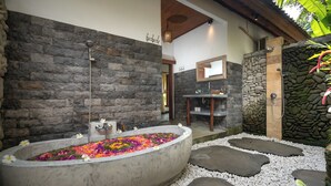 A stone-walled bathroom with a freestanding bathtub filled with colorful flowers, a shower area, and a small wooden table. - Green Sari Private Villa (Ubud)