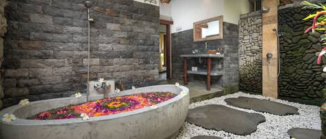 Une salle de bain aux murs de pierre, avec une baignoire sur pieds remplie de fleurs colorées, un espace douche et une petite table en bois.