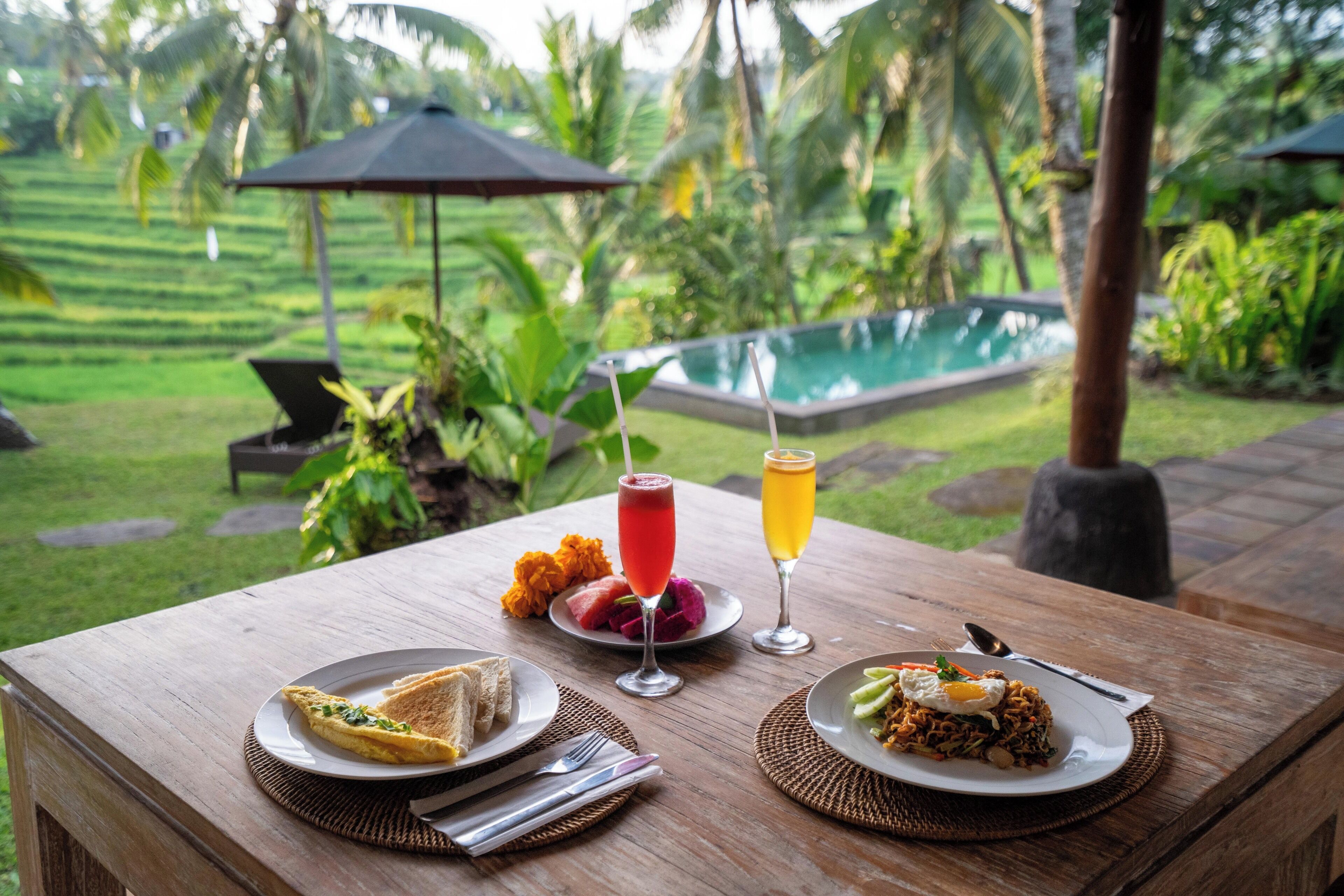 Une table en bois, avec des assiettes de petit-déjeuner, une boisson, et une vue sur une piscine et une végétation luxuriante.