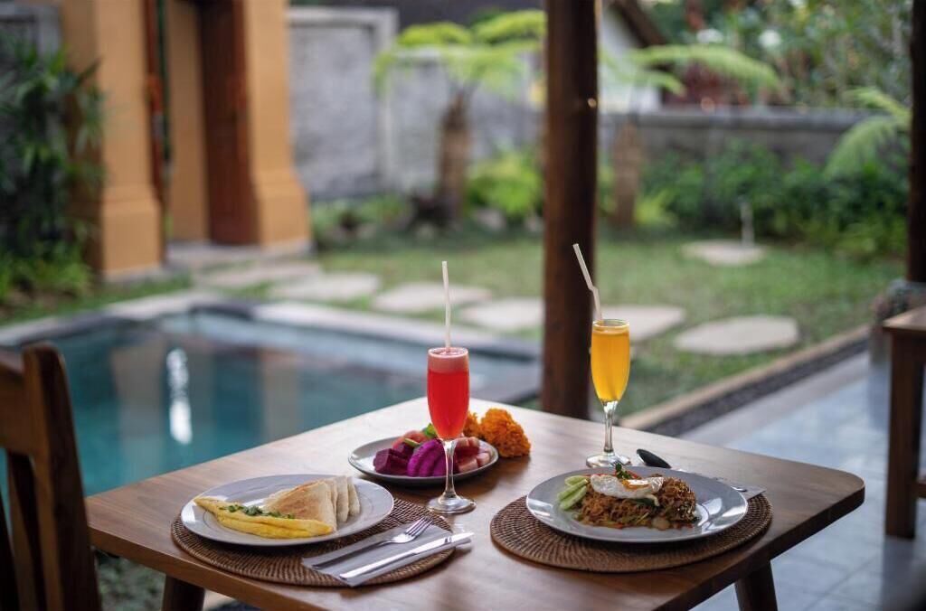 Una mesa de comedor con platos de comida y bebidas, una piscina al fondo y vista a un jardín.