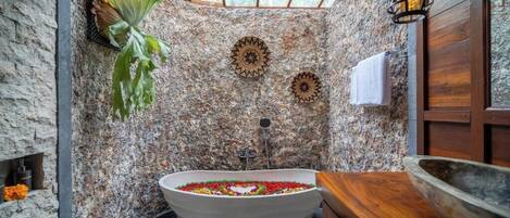 A bathroom with a freestanding bathtub filled with colorful flowers, a stone wall, and a wooden vanity.