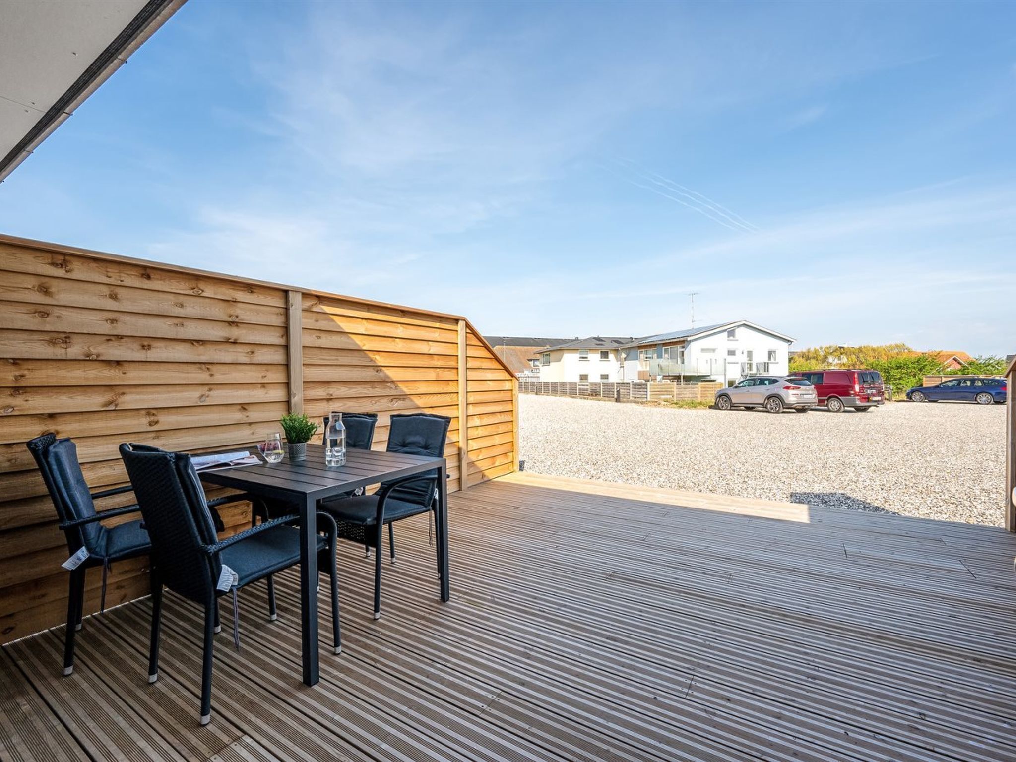 A covered outdoor patio with a table and chairs, a wooden fence, and a gravel driveway leading to a white building.