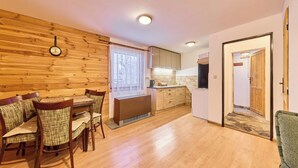 A cozy room with a wooden wall, a dining table with chairs, a window with a view, and a kitchen area with cabinets and a refrigerator. - Chaloupka U lesa by Interhome (Harrachov)