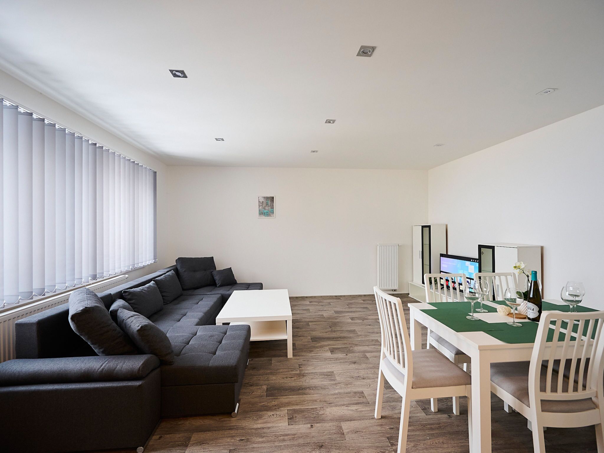 Un salon moderne avec un canapé d’angle, une table à manger avec des chaises et une grande fenêtre équipée de stores.