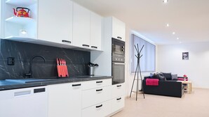 Eine moderne Küche mit weißen Schränken, schwarzen Arbeitsplatten und einem roten Wasserkocher. Im Wohnbereich befindet sich eine schwarz-weiße Couch mit einer rosafarbenen Decke.