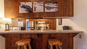 Uma cozinha com armários de madeira, bancada branca, fogão e dois banquinhos de madeira.