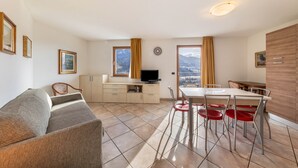 A modern living room with a sofa, dining table, chairs, a TV, and a view of mountains through large windows.