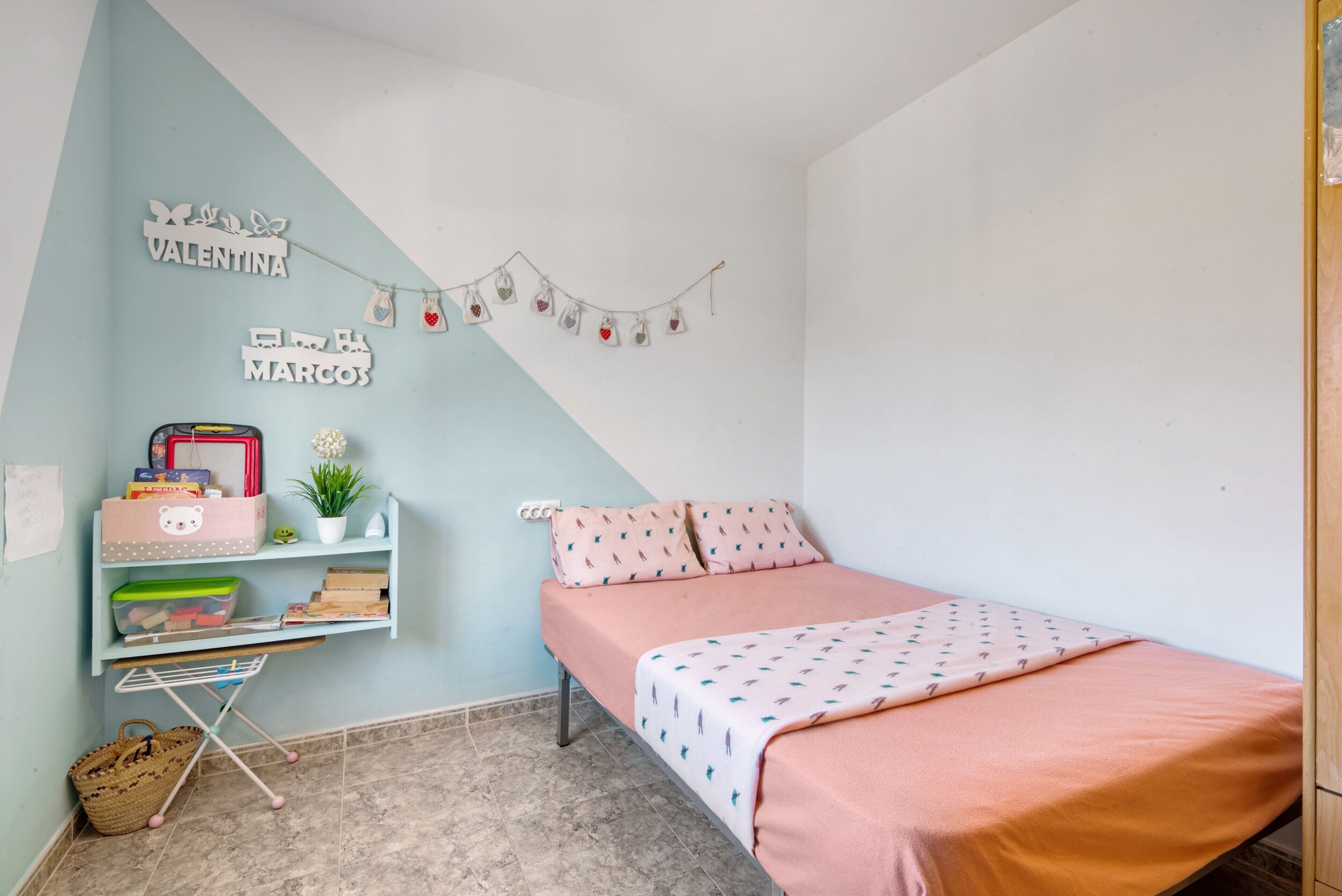Une chambre avec un lit, une étagère remplie de livres et une plante, un mur décoré avec des ornements représentant des noms, et une guirlande d’objets décoratifs en forme de cœur.