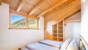 A bedroom with a wooden ceiling, a window with a view of a forest, a bed with pillows, and a wooden wardrobe.
