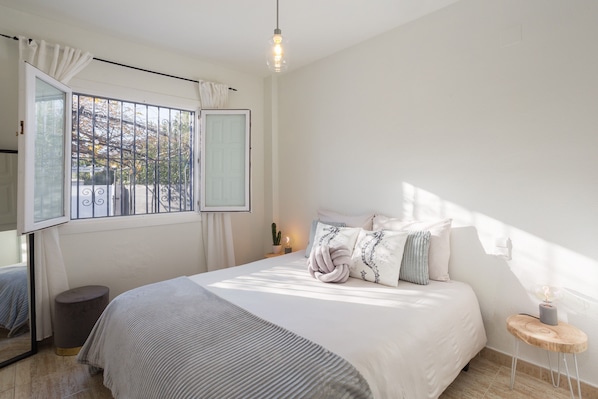 A bedroom with a large bed, a window with curtains, a bedside table with a lamp, and a small stool.
