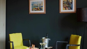 Une pièce avec deux chaises jaunes, une petite table ronde sur laquelle se trouve une boisson, et deux œuvres d’art encadrées accrochées au mur.