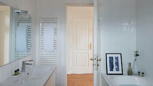 A bathroom with a wooden floor, white walls, a large mirror, a bathtub, and a door leading to another room.
