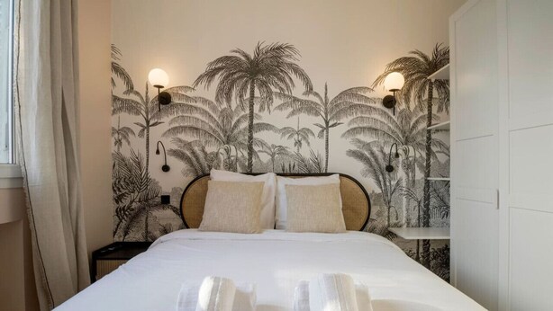 A hotel room with a bed, white bedding, a headboard with a palm tree mural, two wall-mounted lamps, and a wardrobe. - 11.Appart 4Pers#1 Bedroom #Père Lachaise #Nation (Paris)