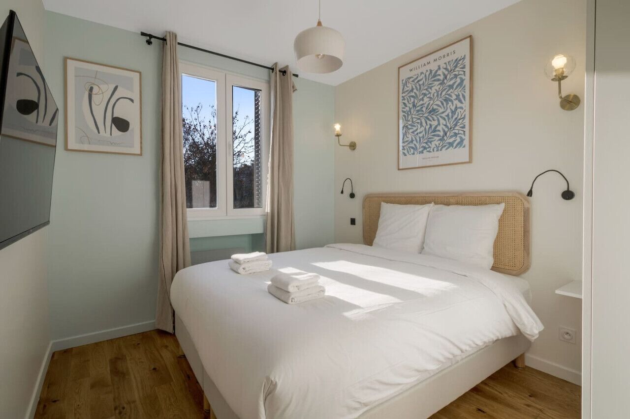 Un dormitorio con una cama grande, una ventana con cortinas, lámparas fijadas en la pared y cuadros enmarcados.