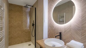 Ein modernes Badezimmer mit Wandpaneelen in Fischgrätmuster, einem runden Spiegel mit integrierter Beleuchtung, einem weißen Waschbecken und einer Duschkabine aus Glas.