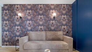 Ein Wohnzimmer mit gemustertem Wandpapier, einem beigen Sofa, zwei Wandleuchten und einer kleinen weißen Vase auf einem Holztisch.
