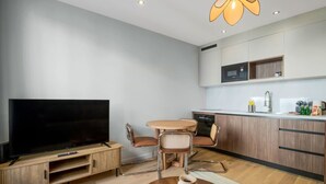 Ein modernes Wohnzimmer mit Flachbildfernseher, Holz-Entertainment-Möbel, rundem Esstisch mit Stühlen und einer Küchenzeile mit Schränken und Mikrowelle.