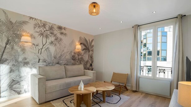 Un salon moderne avec un canapé beige, une table basse en bois et une fresque représentant un feuillage tropical sur le mur.