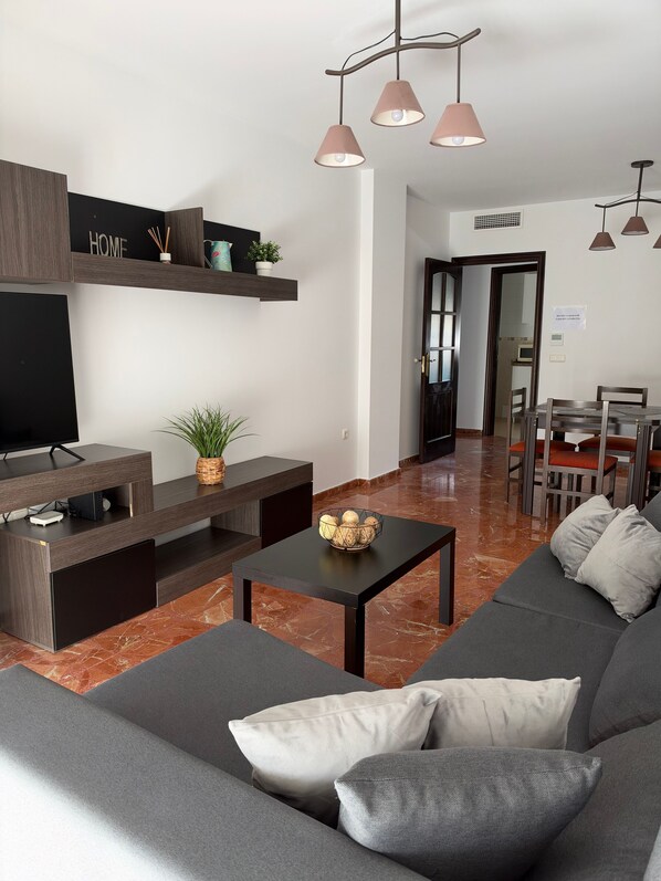 A modern living room with a sofa, coffee table, TV stand, and a dining area with chairs and a table.