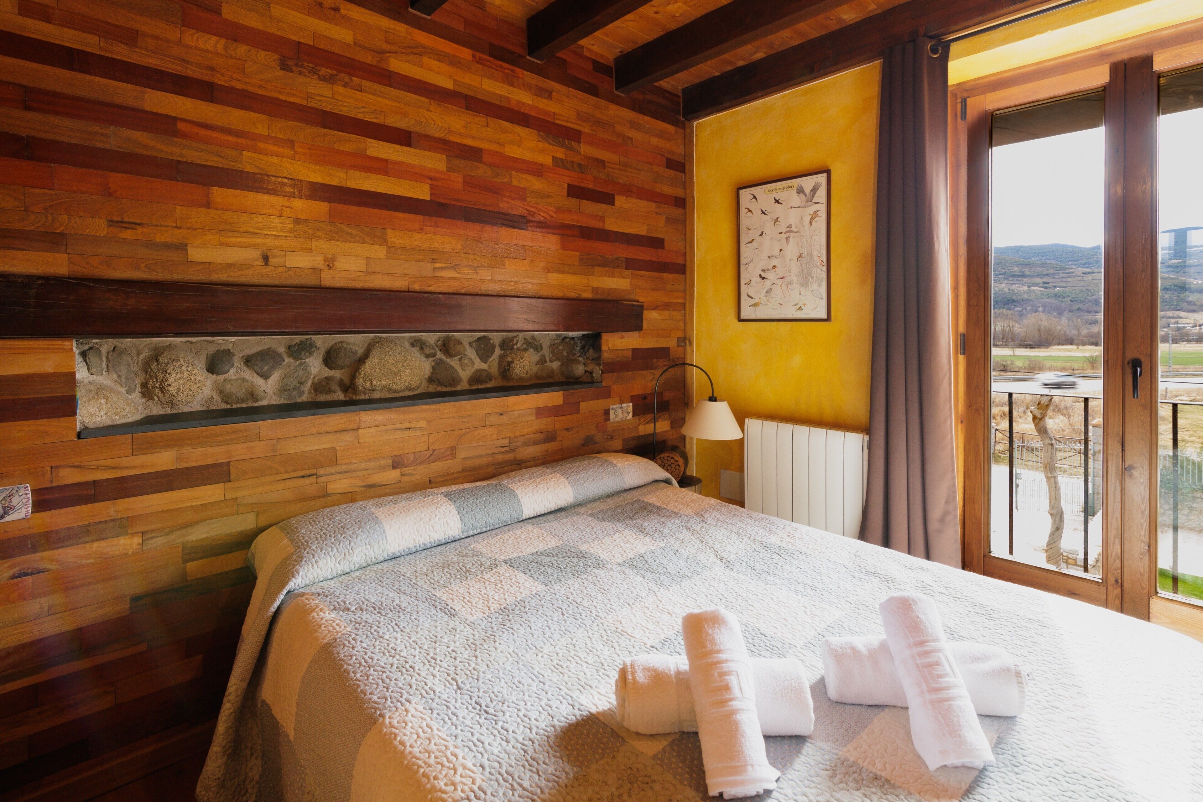 Une chambre avec un mur en bois, une étagère en pierre, un lit recouvert d’une couverture à carreaux et une vue sur un pont et un paysage, visible par une fenêtre.
