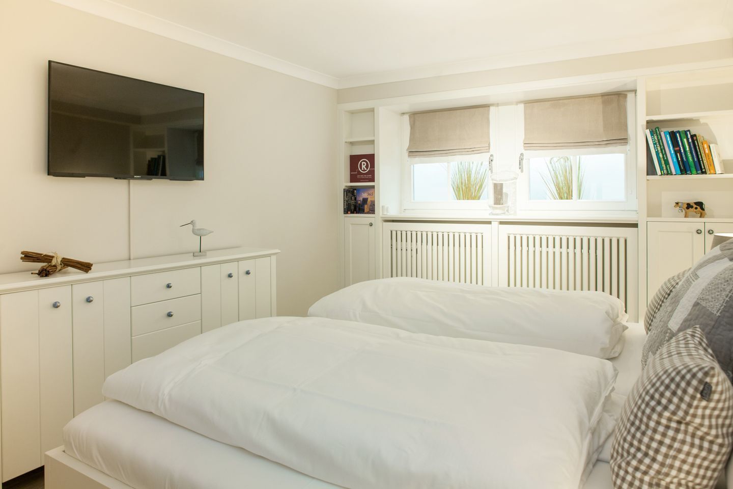 A bedroom with a bed, a TV mounted on the wall, a radiator, and a bookshelf with books.
