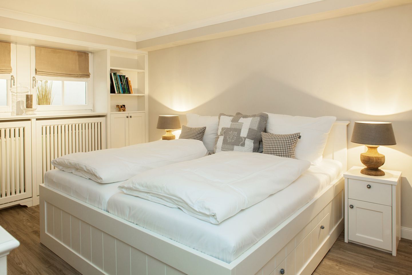 A neatly made bed with white bedding, two bedside tables with lamps, a bookshelf, and a radiator.