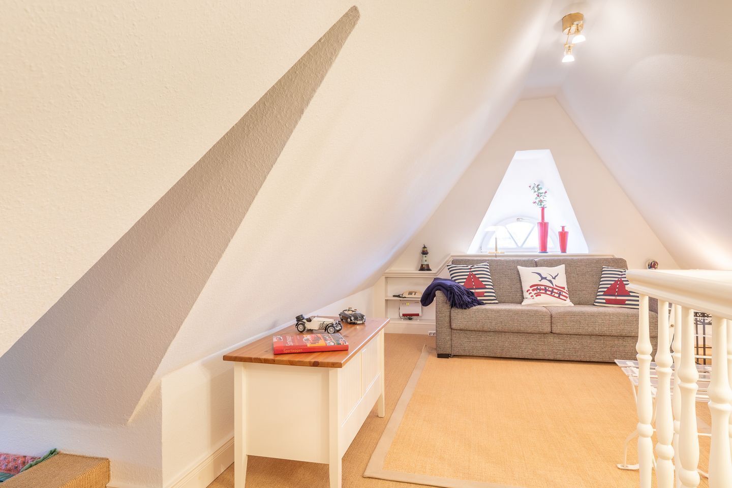 A cozy attic bedroom with a sloped ceiling, a sofa with nautical-themed pillows, a small table with toys, and a window with a view of a sailboat.
