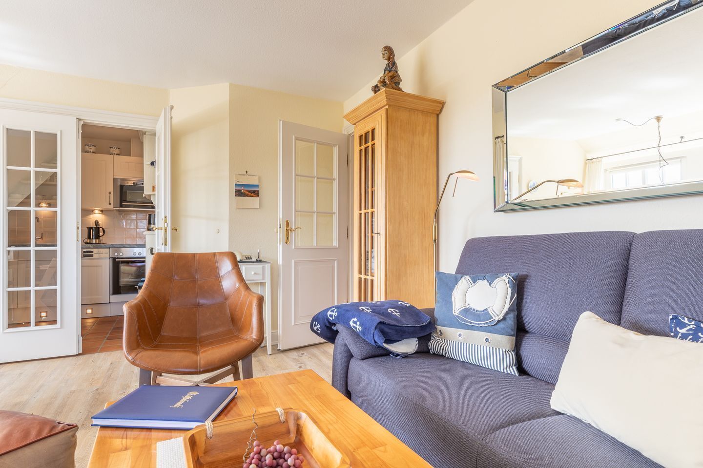 Ein Wohnzimmer mit einer Couch, einem Holztisch, einem Ledersessel und einer durchgehenden Tür erkennbaren Küche.