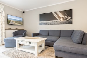 Un salon avec un canapé gris, une table basse blanche, un téléviseur à écran plat et un tableau représentant un voilier.