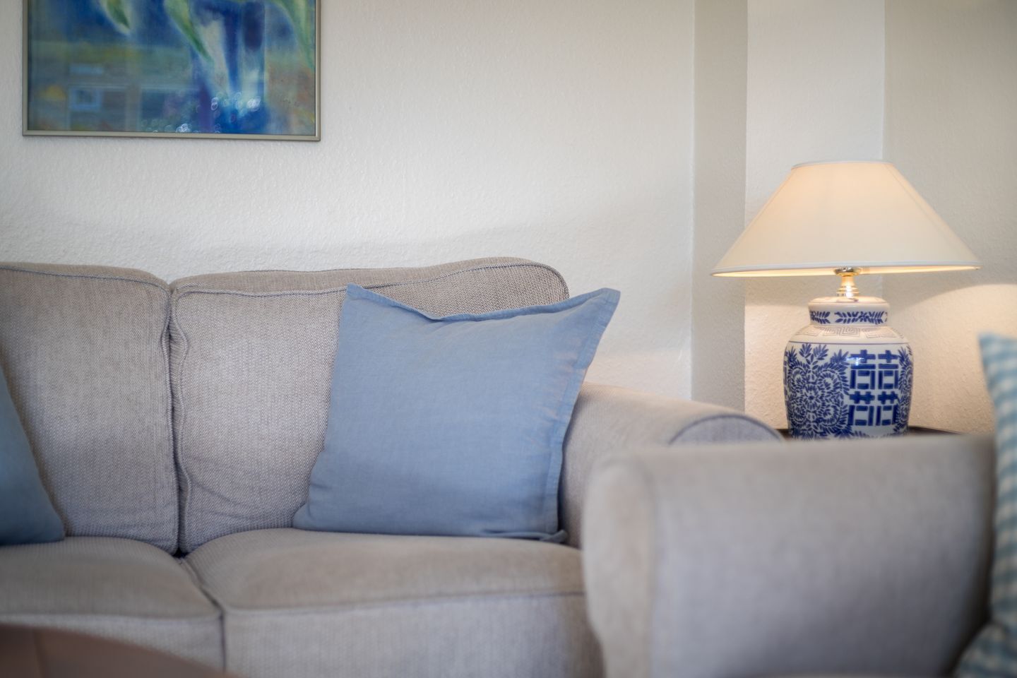 Un canapé beige avec un coussin bleu, une lampe bleue et blanche, et un tableau accroché au mur.