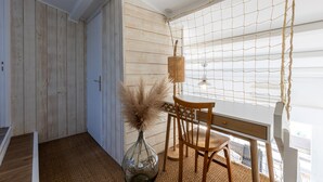 Een kamer met een houten vloer, een witte deur, een rieten stoel, een houten tafel en een raam met een witte zonwering.