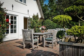 A white house with a patio area featuring a wooden table and chairs, surrounded by greenery and a wooden fence.