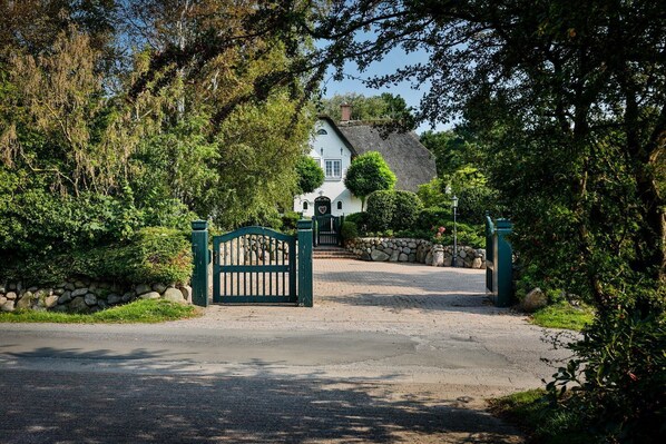 A gated entrance to a white building with a thatched roof, surrounded by lush greenery.