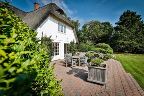 A white building with a thatched roof, a patio with wooden furniture, and a well-maintained garden.