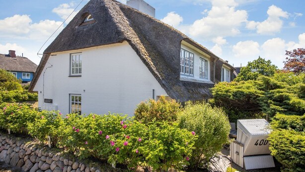 Une maison traditionnelle à toit de chaume, aux murs blancs, entourée d’une végétation luxuriante et d’un mur de pierre.
