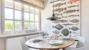 Un coin repas avec une table ronde dressée pour quatre personnes, des chaises blanches, un radiateur, une fenêtre donnant sur un paysage verdoyant et une illustration de poissons accrochée au mur.