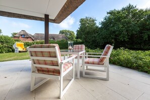 Eine Terrasse mit zwei weißen Stühlen mit karierten Kissen, einem Glastisch und einem überdachten Strohdach.