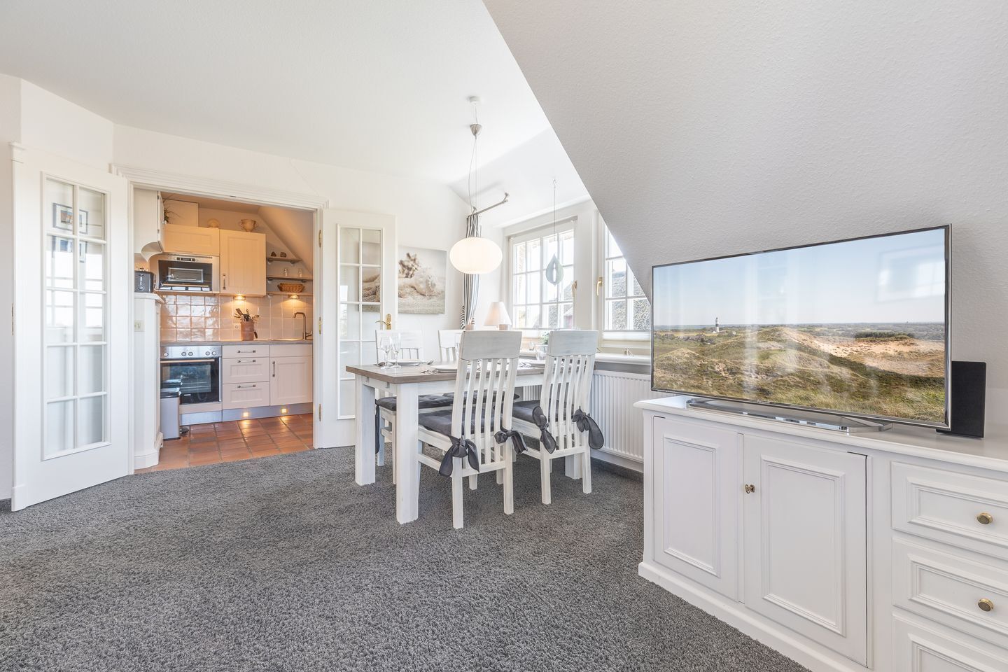 A modern kitchen with white cabinetry, a dining area with a table and chairs, and a large TV displaying a coastal landscape.