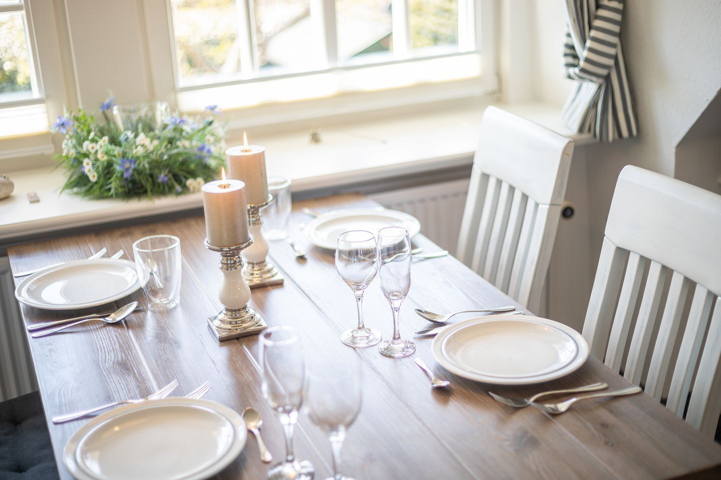 Une table à manger dressée avec de l’argenterie, des assiettes blanches et des verres à vin, entourée de chaises blanches.