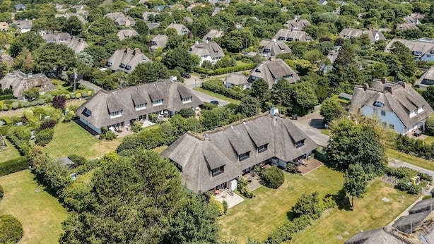An aerial view of a residential area with houses featuring thatched roofs, surrounded by greenery and a body of water in the background. - Sharky Bay - Die Reetgedeckte, Gepflegte Anlage Liegt auf Einem ca. 2000 m² Großem Grundstück (Sylt-Ost)