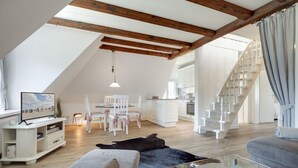 A spacious loft with a high ceiling, exposed wooden beams, a dining area, a kitchen, and a staircase. - Sharky Bay - Die Reetgedeckte, Gepflegte Anlage Liegt auf Einem ca. 2000 m² Großem Grundstück (Sylt-Ost)
