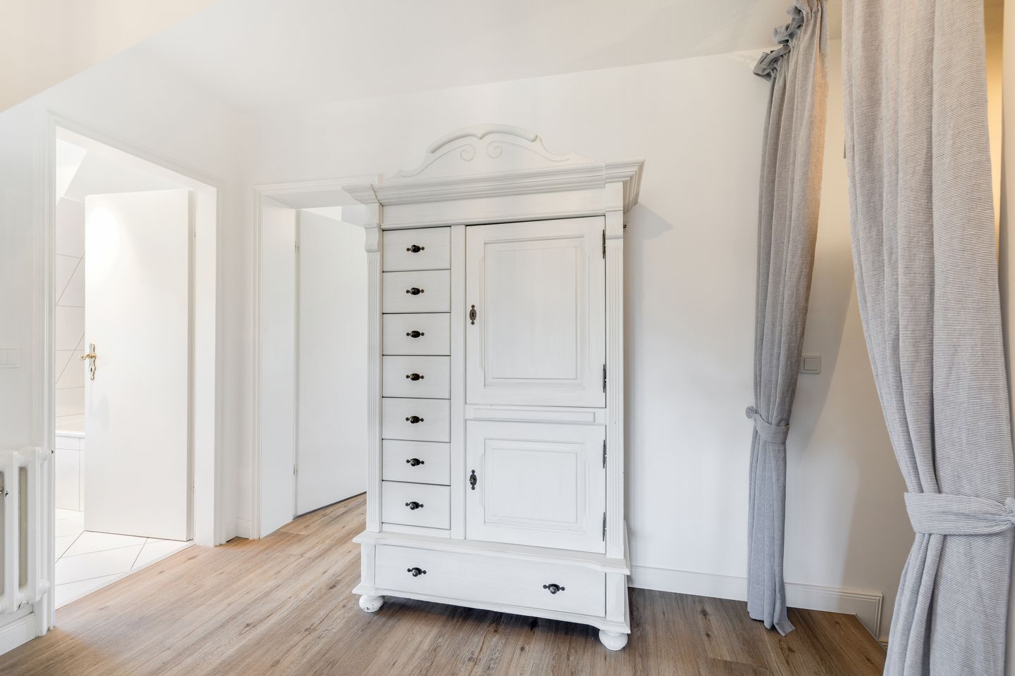Un armario blanco con varios cajones y puertas, piso de madera y una puerta que da a otra habitación.