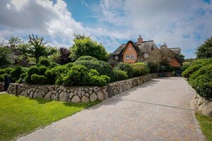 A stone pathway leading to a traditional house with a thatched roof, surrounded by well-maintained hedges and a stone wall.