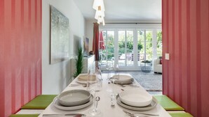 Spisestue med et langt bord dækket til fire personer, en moderne pendellampe og udsigt gennem en glasdør til en udendørs patio med en hvid stol og et lille bord.
