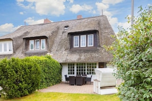 Et traditionelt hus med stråtækt tag, en terrasse med havemøbler og en velplejet have.