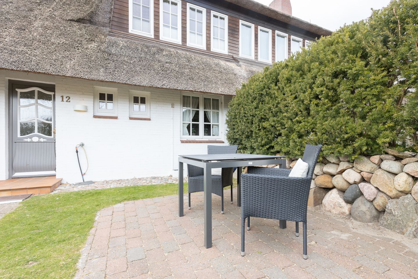 A house with a thatched roof, white exterior walls, and a number '12' on the door. There is a patio with a table and chairs, and a well-maintained hedge to the right.